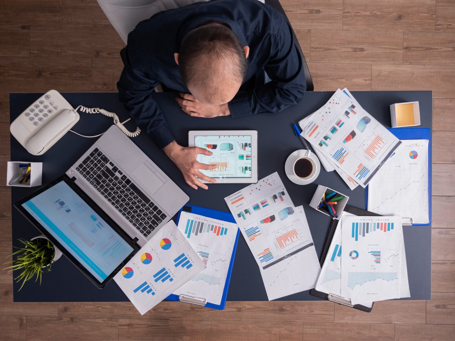 High-angle overhead view of a professional sitting at an office desk surrounded by financial reports, colorful data graphs, paperwork, an open laptop, and a digital tablet.